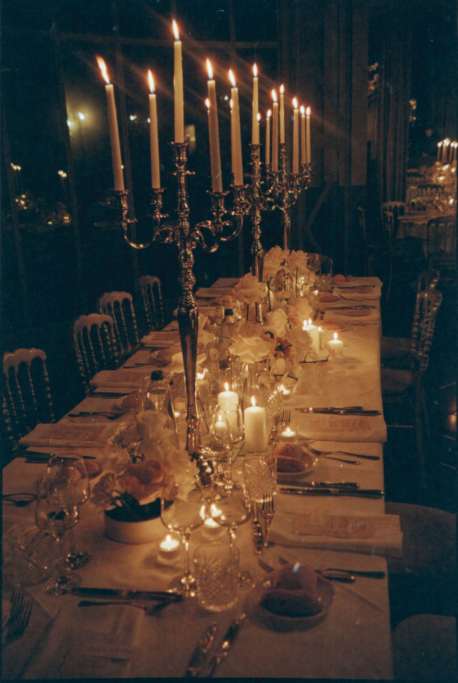 Long candlelit dining table set for an elegant dinner party with glowing mason jar centerpieces in a dark room.