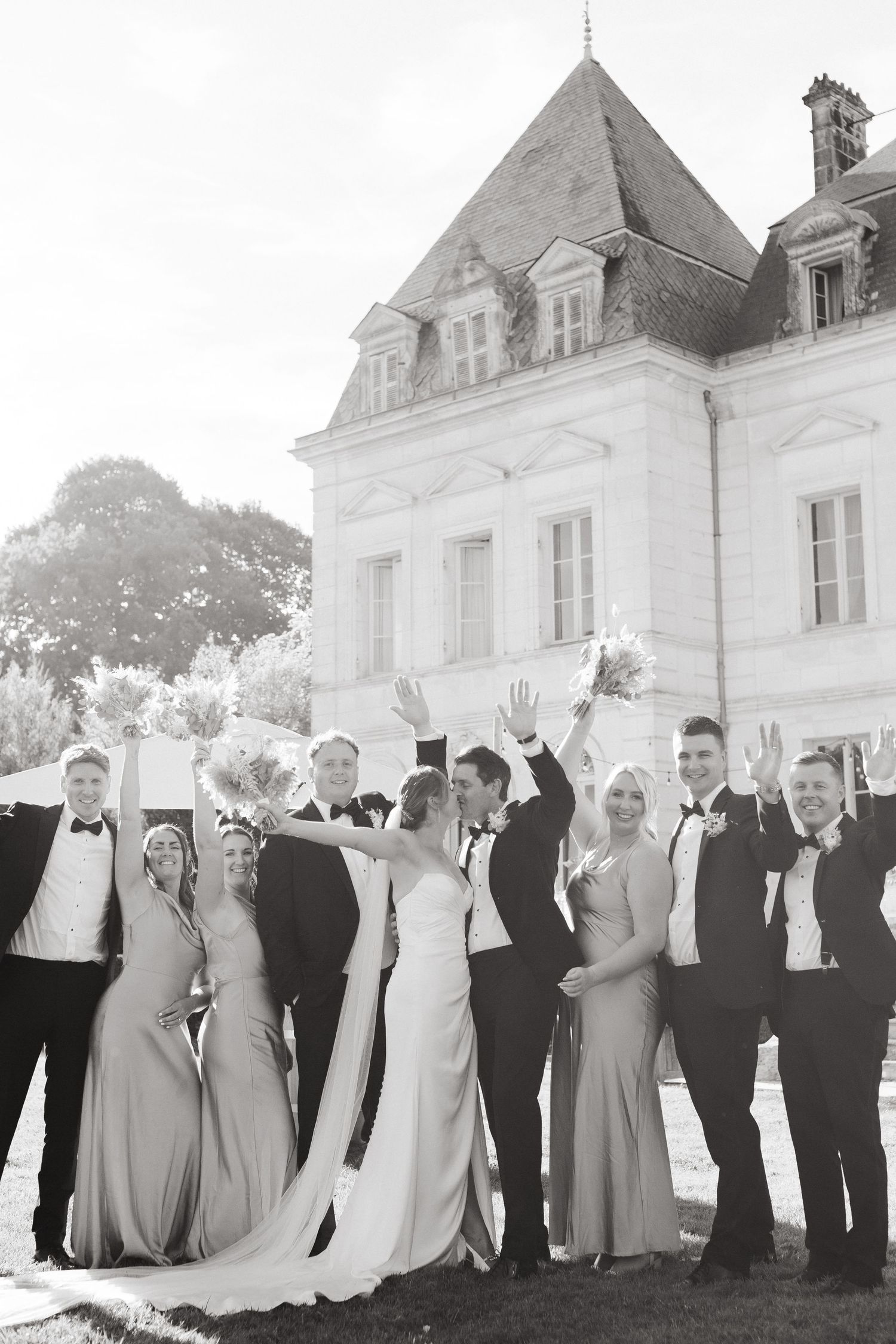 Black and white panoramic shot of a wedding party lined up outdoors with guests raising their hands in celebration.