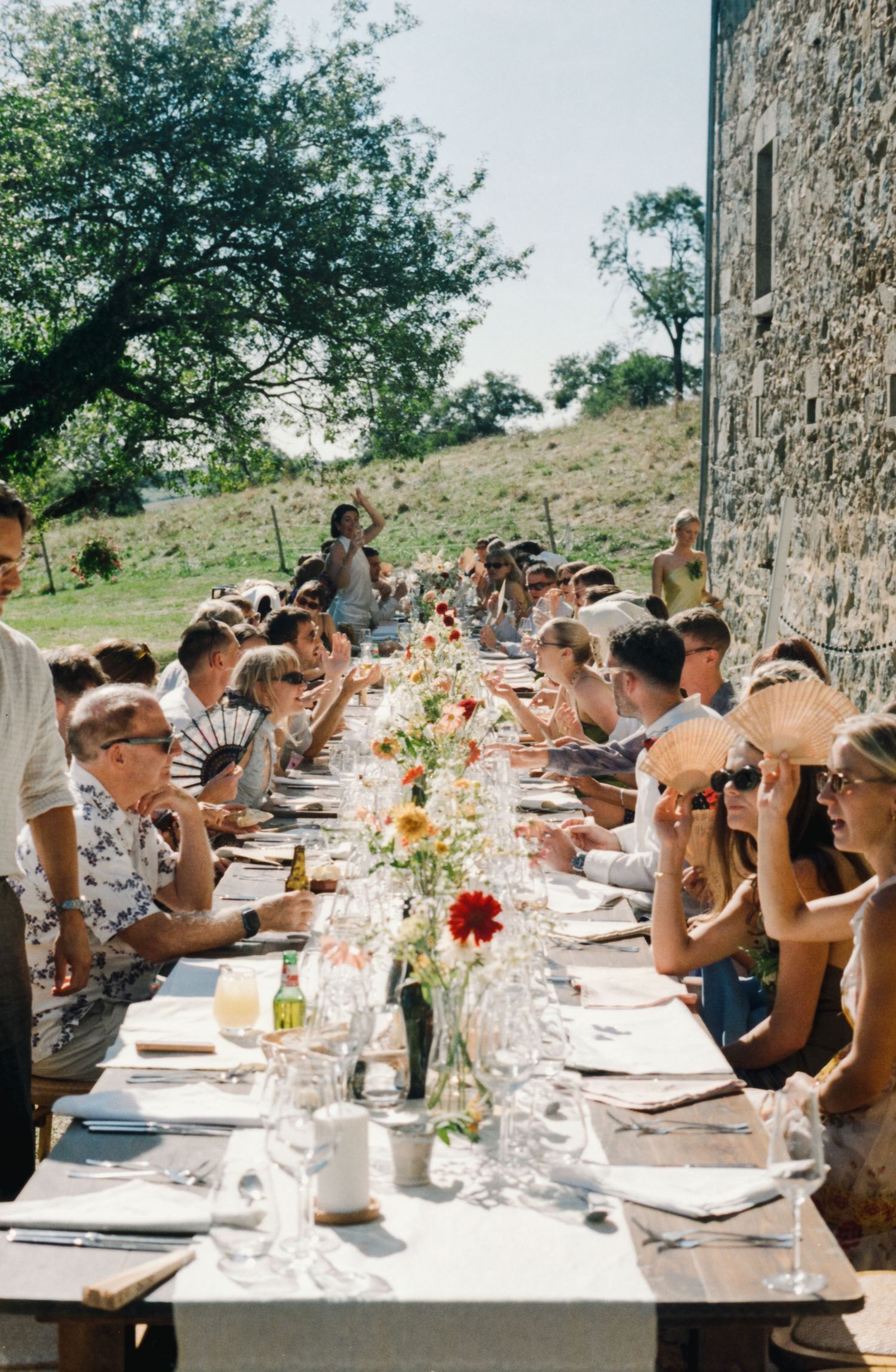 A long outdoor dining table set for a summer celebration with white linens and floral centerpieces against a stone wall.