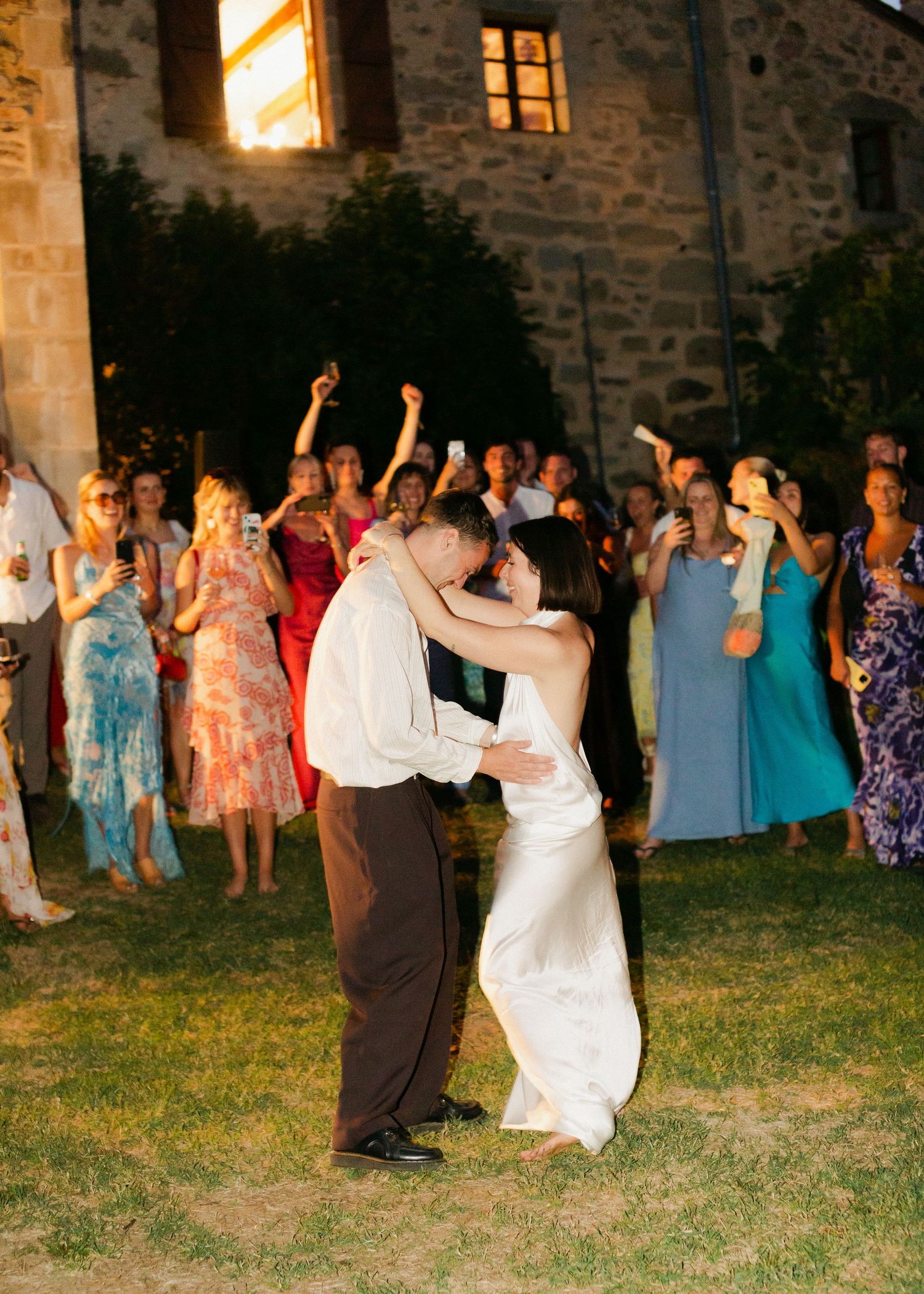 Wedding guests dance and celebrate on a grassy lawn outside a rustic stone building at dusk.