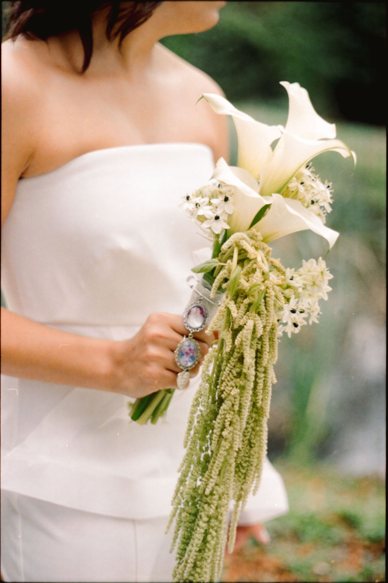 A delicate white floral bouquet with cascading greenery is held against a white wedding dress in a garden setting.