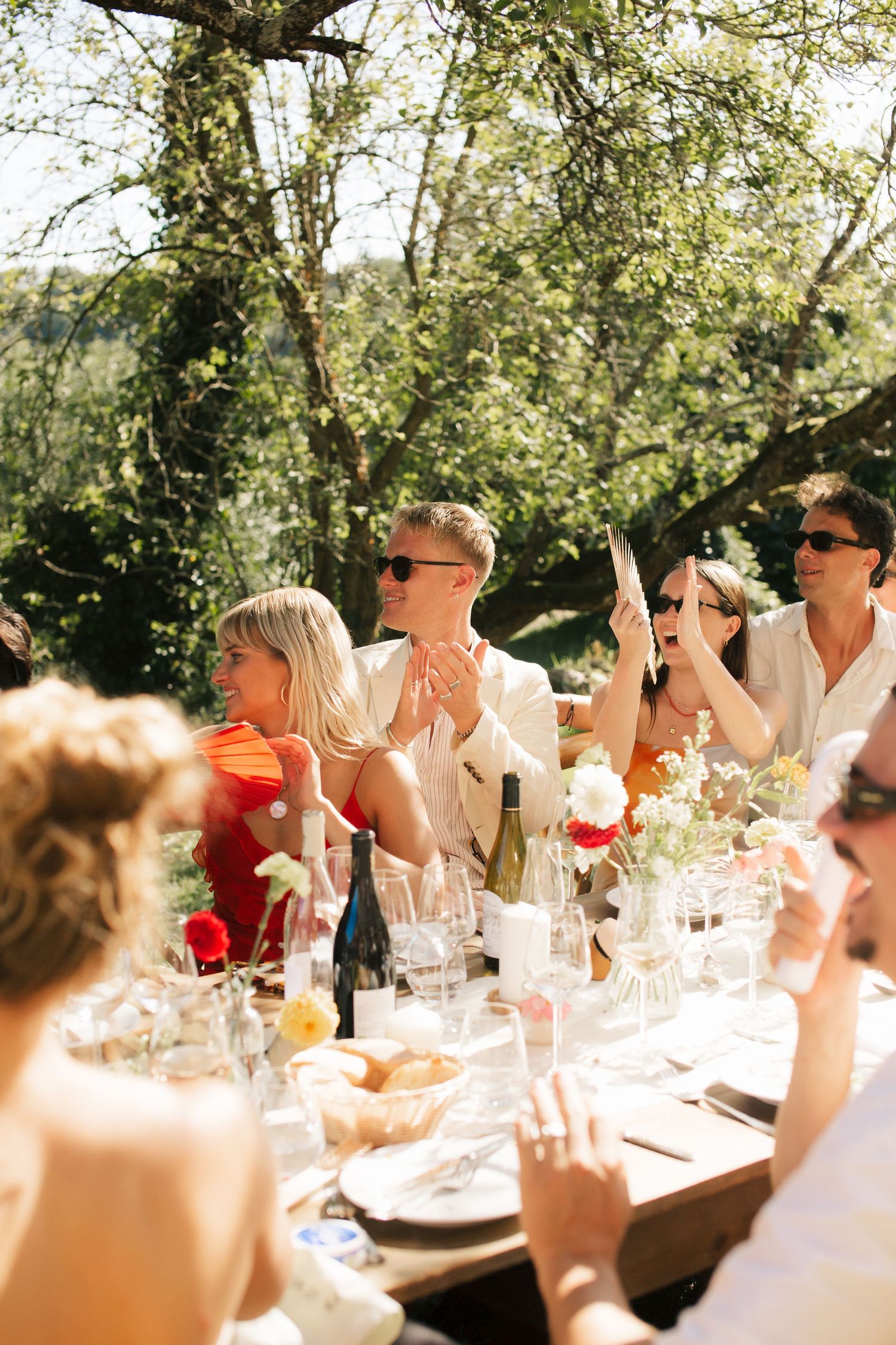 Wedding guests sit at an elegant outdoor reception table under trees in warm sunlight during a summer celebration.