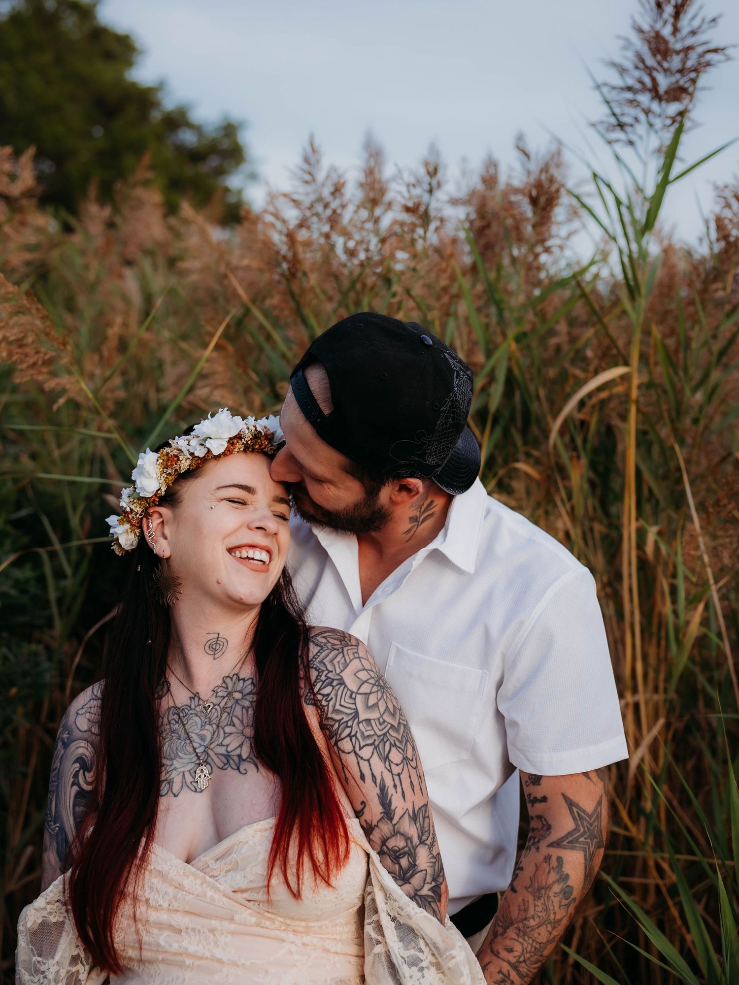 Heavily tattooed couple laughing together in tall grass, woman wearing floral crown and lace dress.