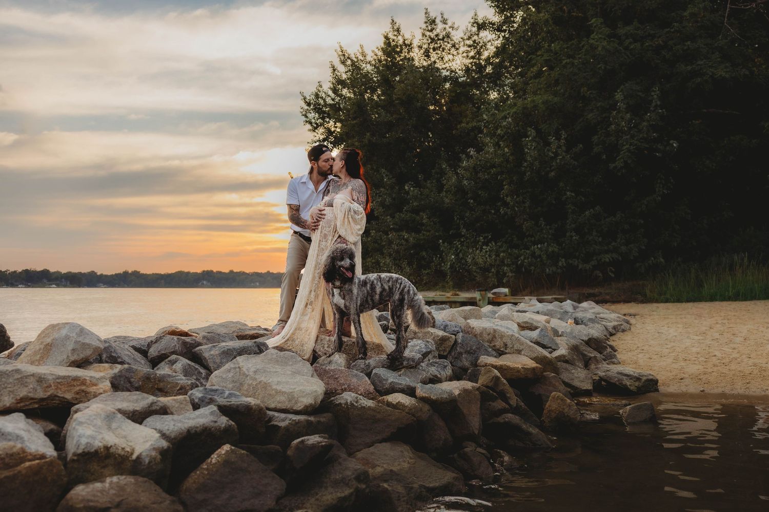 Couple embracing on rocky shore at sunset with large dog, woman wearing flowing lace maternity dress.