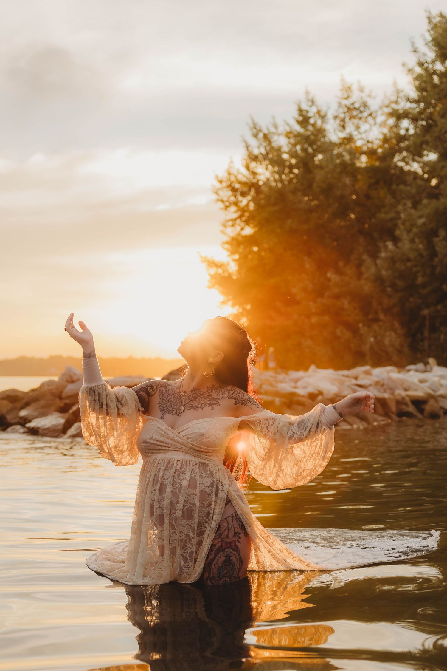 Pregnant woman in lace dress standing in water at golden sunset with arms outstretched joyfully.