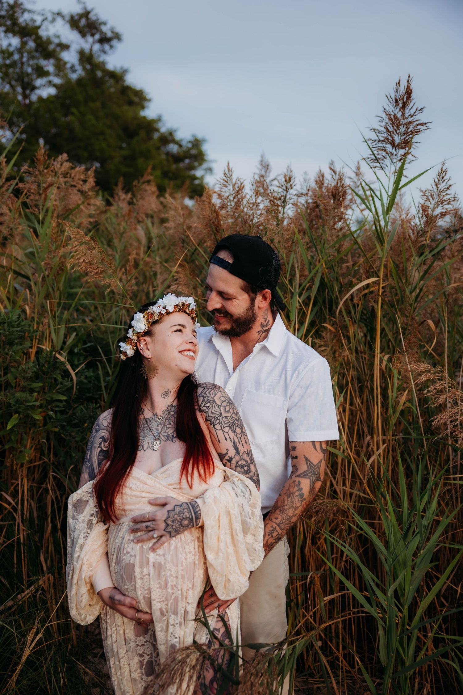 A tattooed expectant couple embraces tenderly among tall golden reeds at dusk.