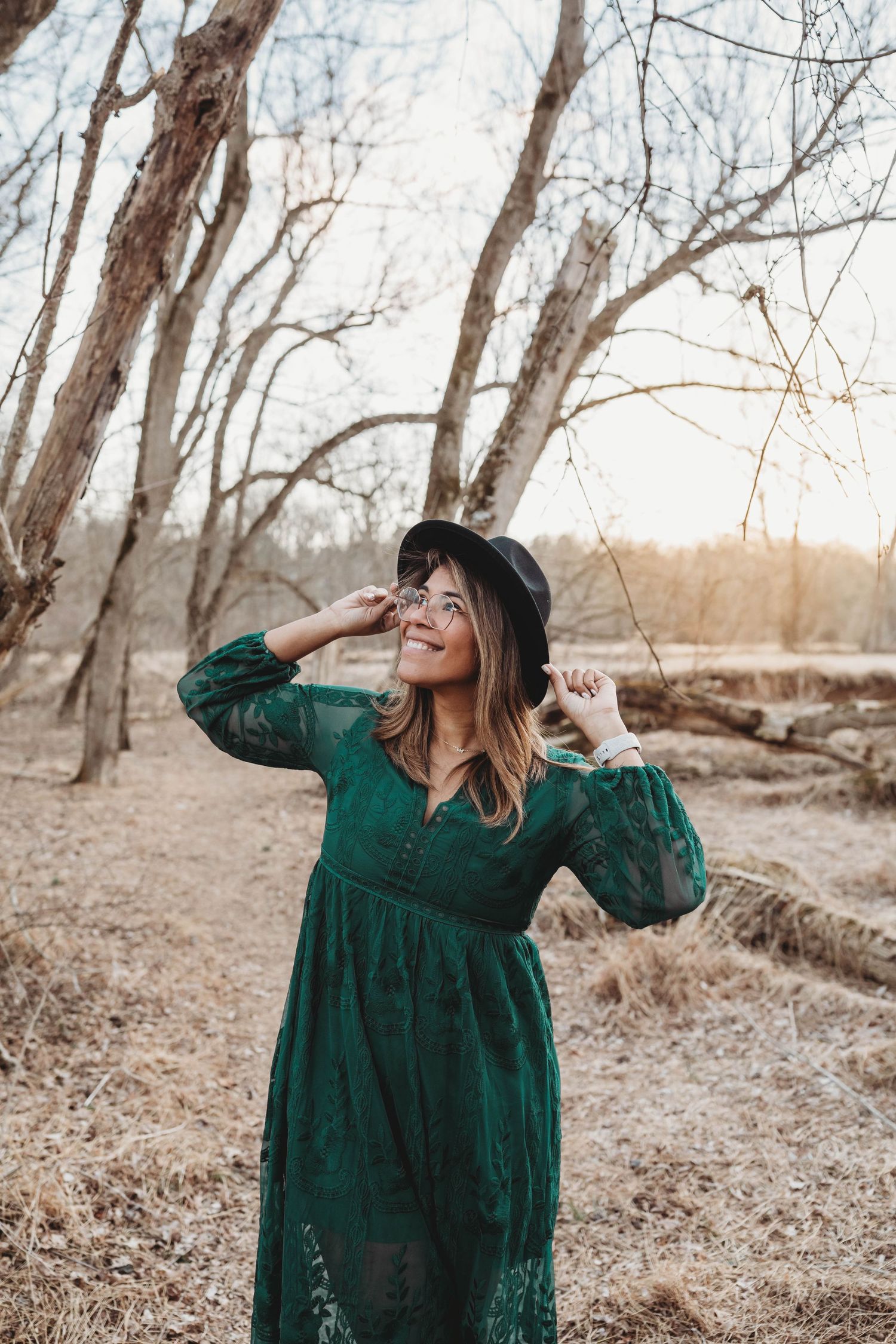 A smiling woman in a green lace dress and black hat poses among bare trees at golden hour.