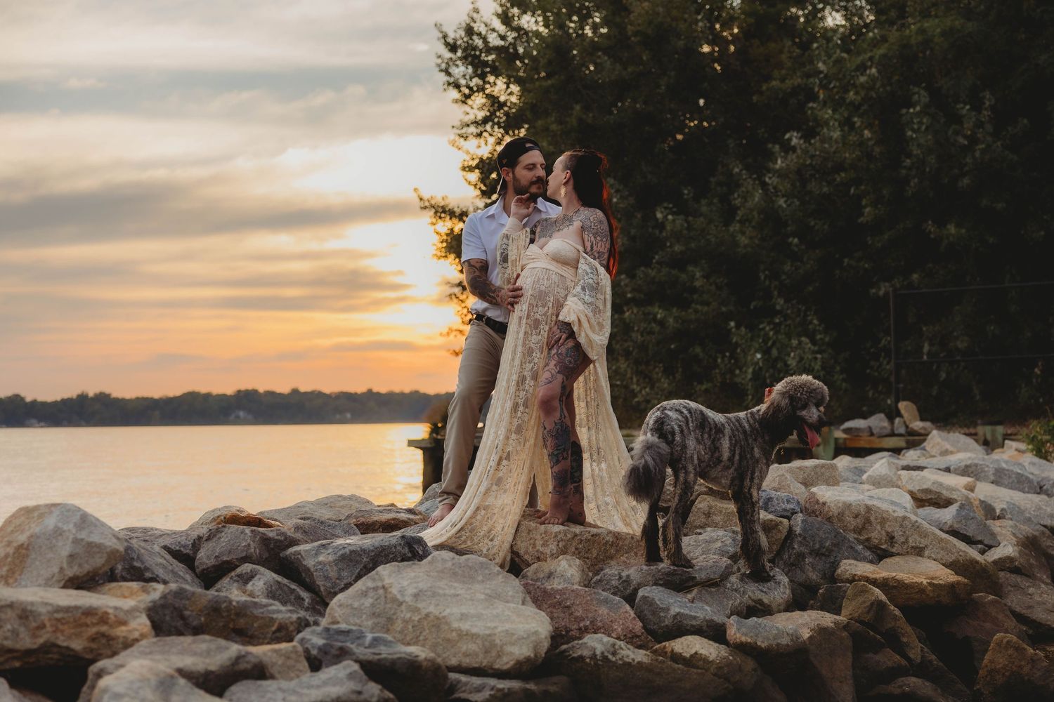 Tattooed couple with dog posing on rocky waterfront at sunset, woman in elegant lace maternity gown.