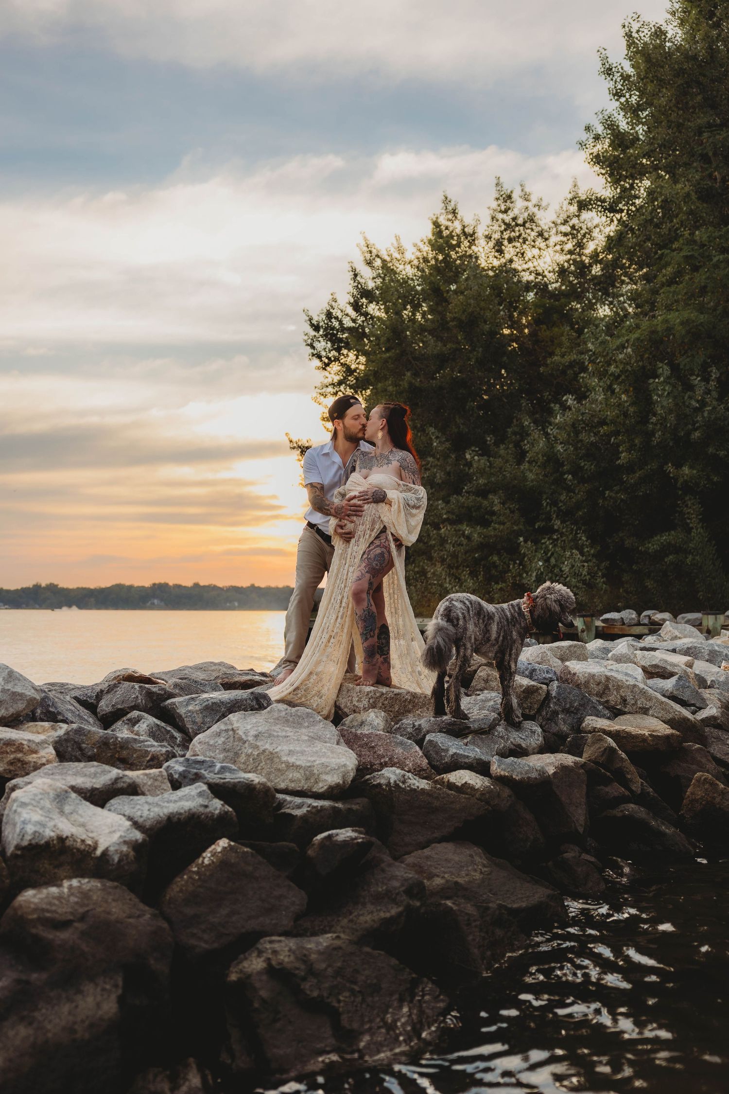 Couple with tattoos and dog on rocky shore at sunset, pregnant woman in lace dress embraced by partner.