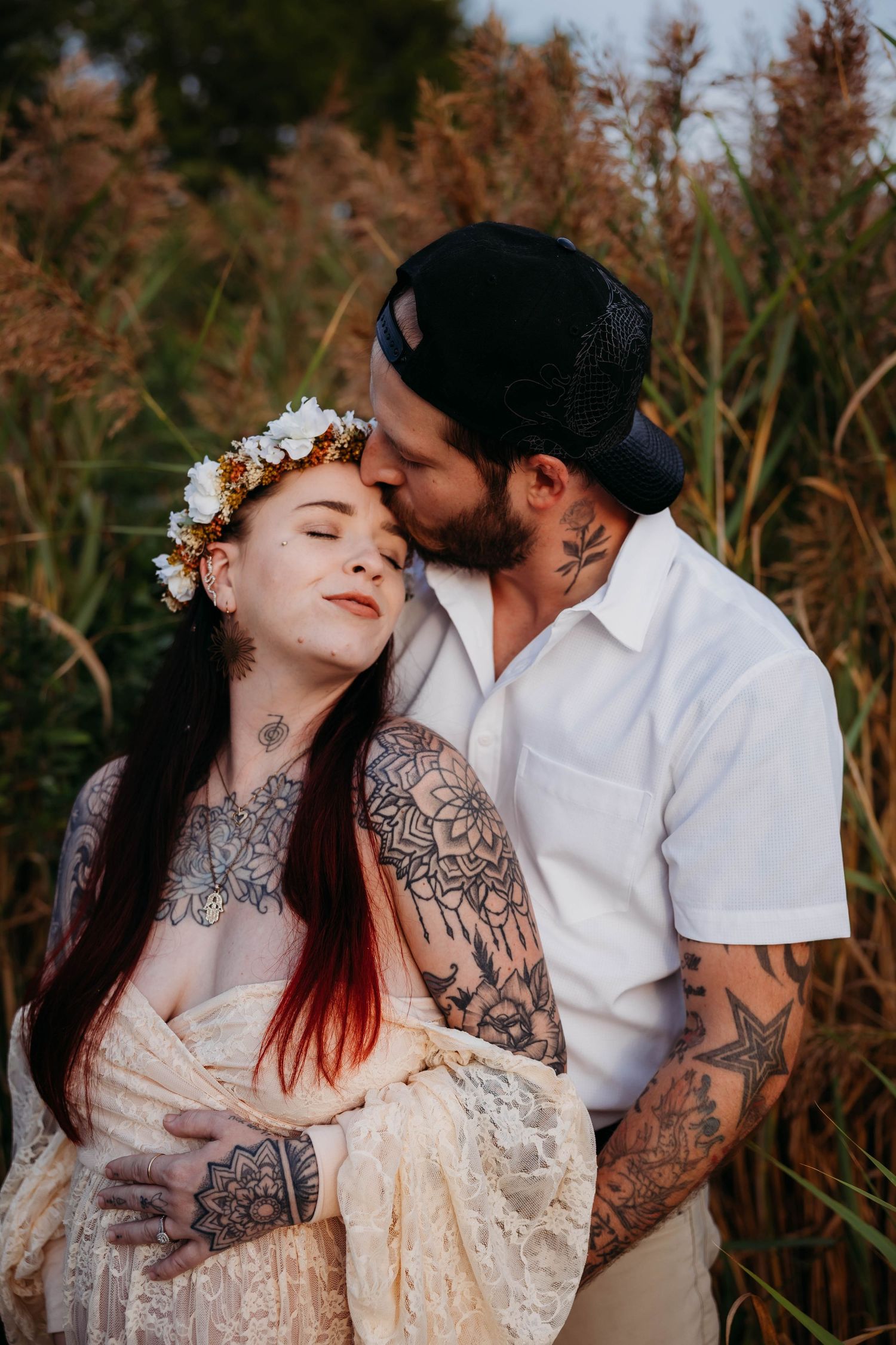 Tattooed couple sharing tender kiss in golden field, woman in floral crown and lace maternity dress.