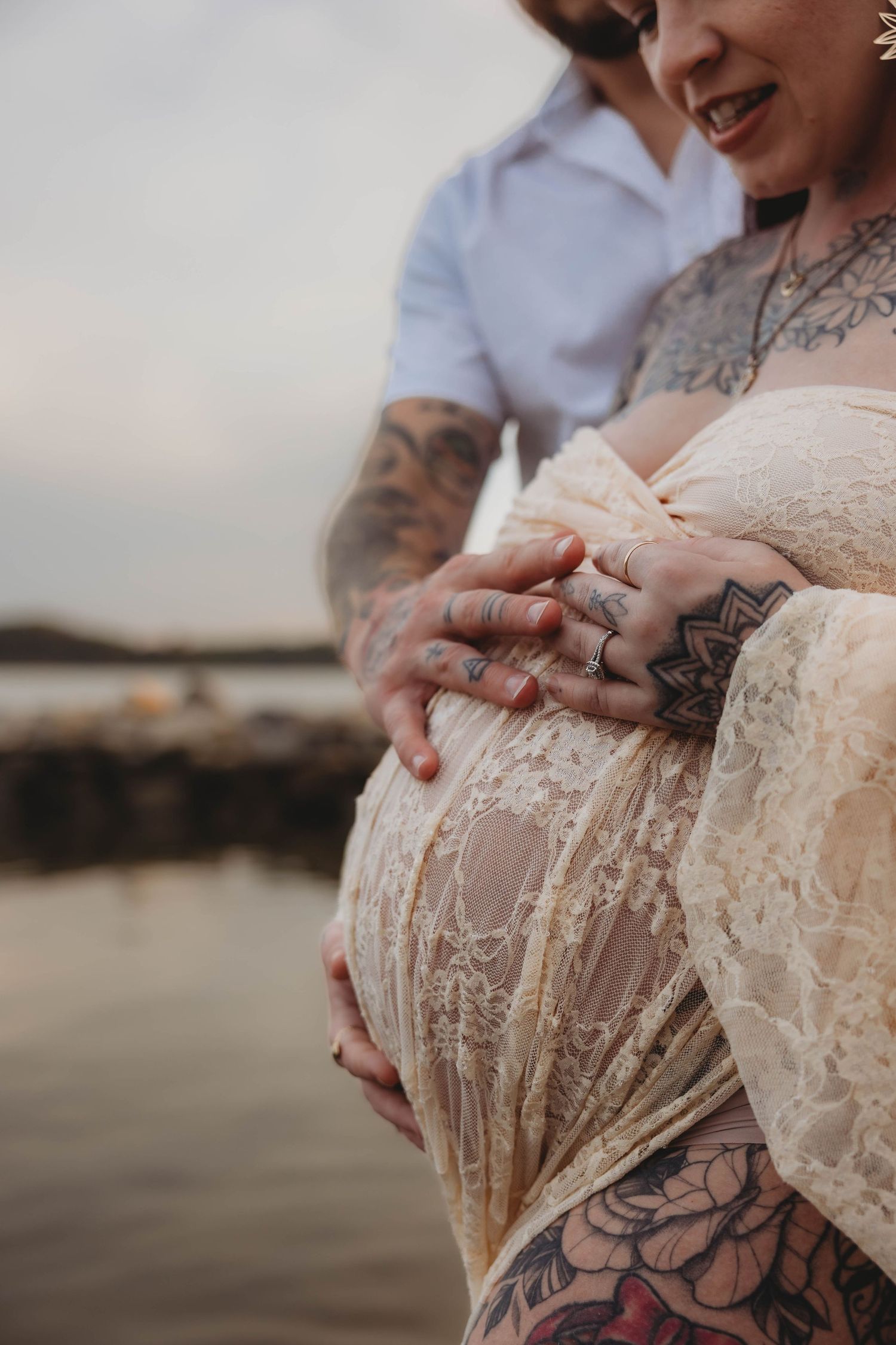 Close-up of tattooed couple cradling pregnant belly in sheer lace maternity dress near water.
