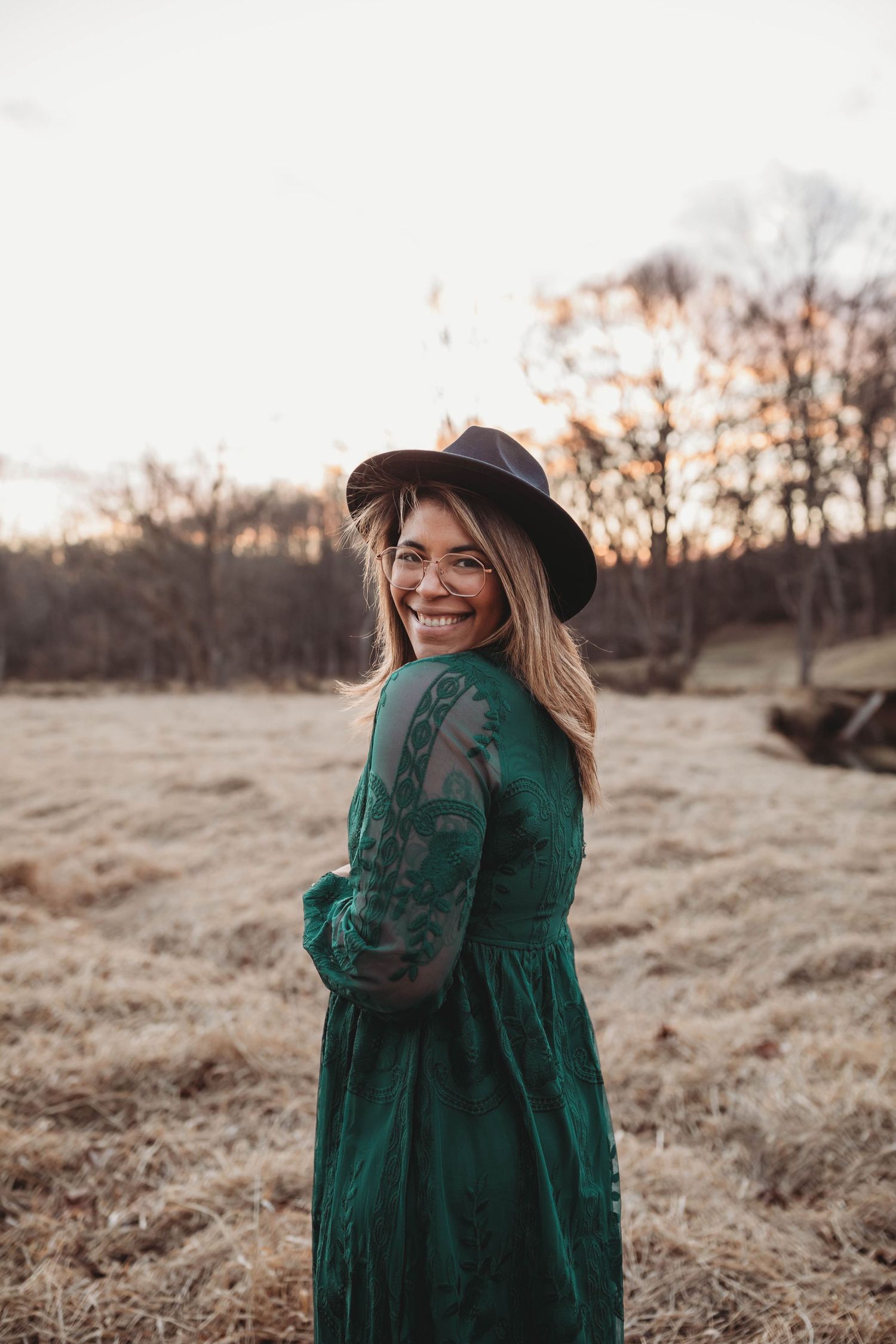 A laughing woman in a green embroidered dress and black hat stands in a golden autumn field.