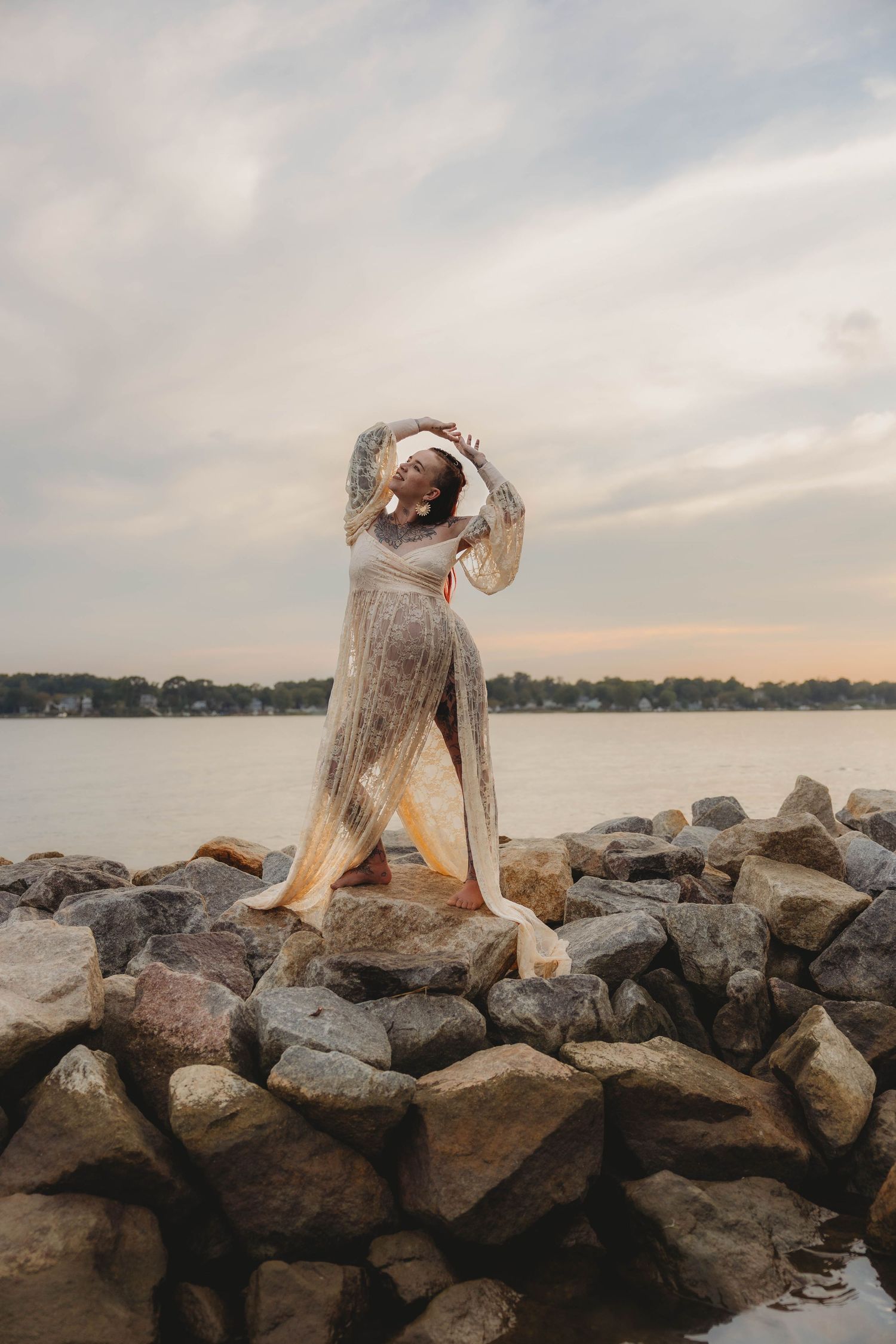 Tattooed pregnant woman in flowing lace dress posing gracefully on rocky shoreline at dusk.