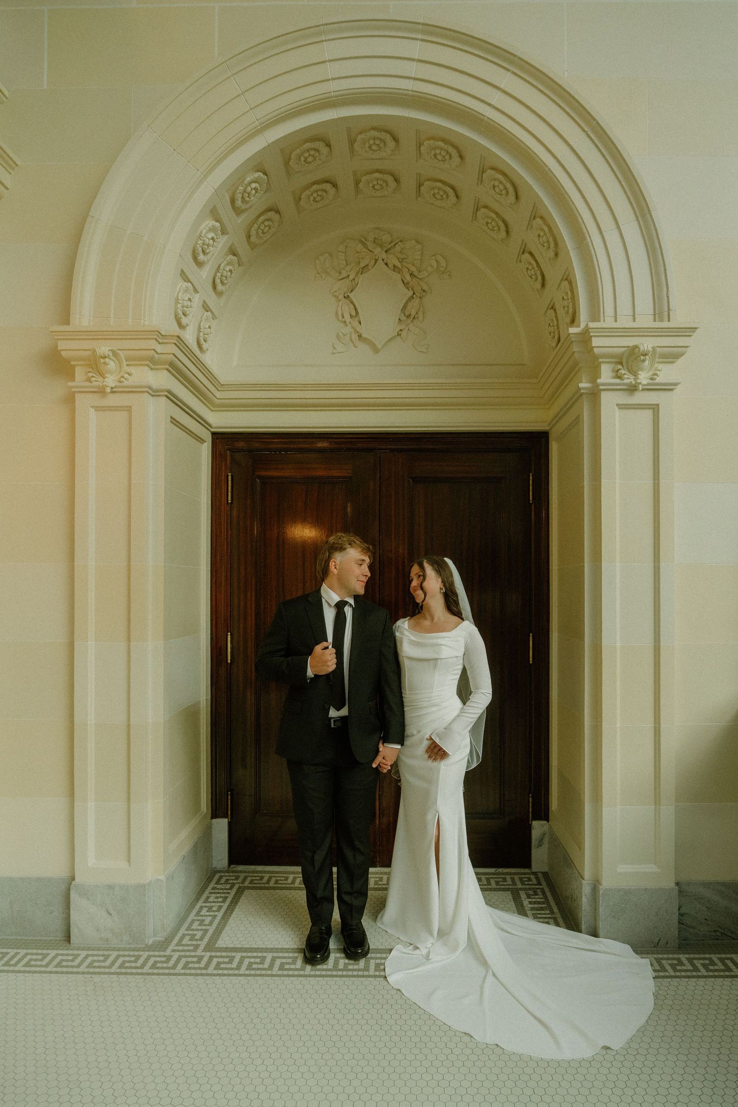 Wedding couple in elegant white dress and suit pose under arched doorways in a classic architectural setting.