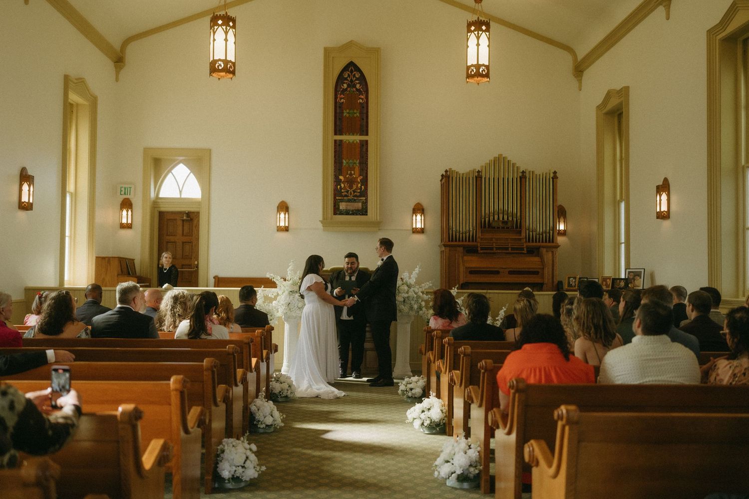 A traditional wedding ceremony takes place in a small chapel with wooden pews and stained glass windows.