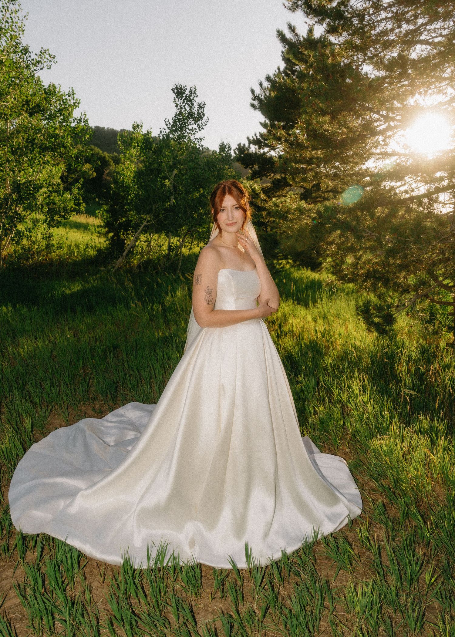 A flowing white wedding dress billows gracefully across green grass as golden sunlight filters through trees at sunset.