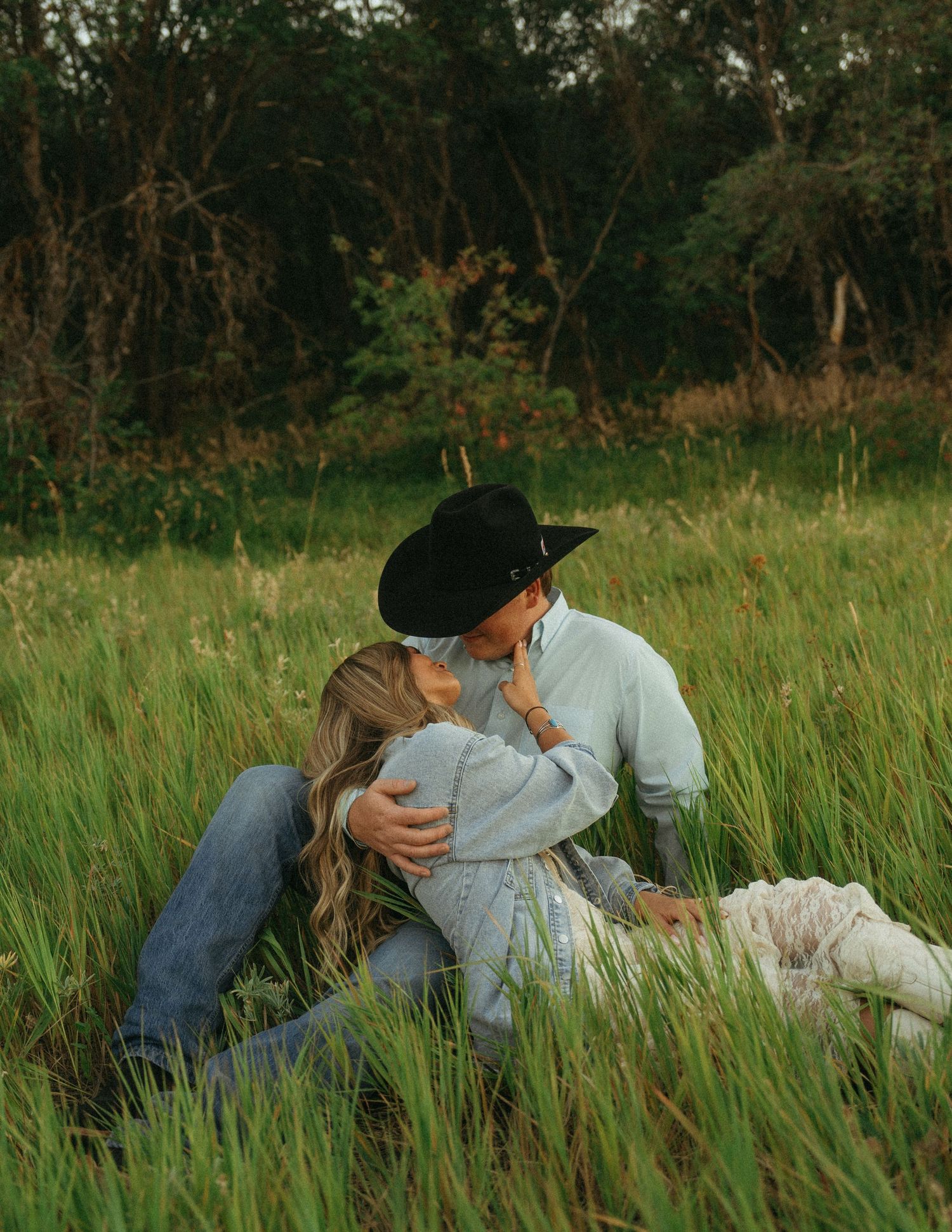A couple in western attire sharing an intimate moment while sitting in tall grass at sunset.