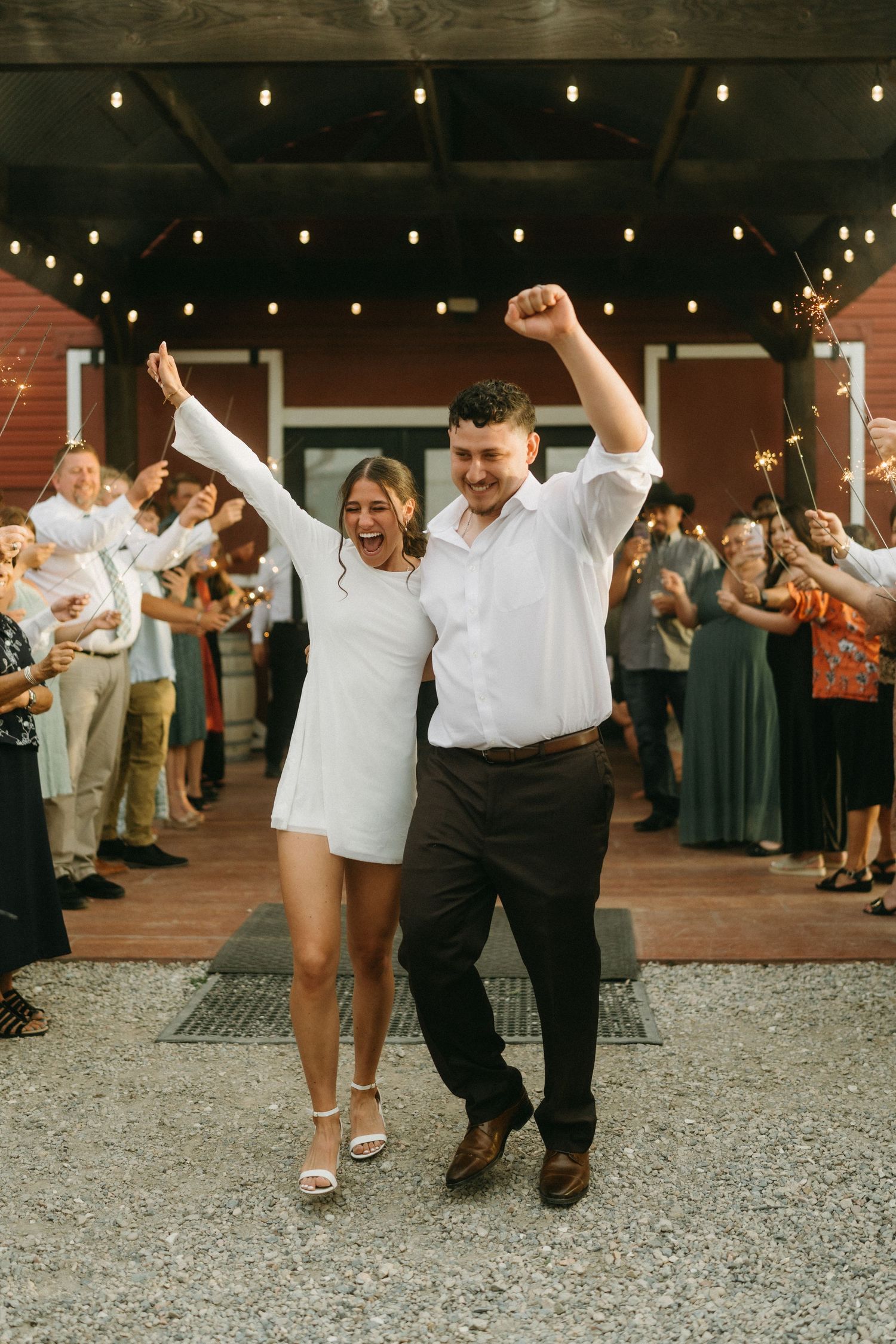 Wedding guests dance in a line on a gravel patio under string lights at an outdoor reception celebration.