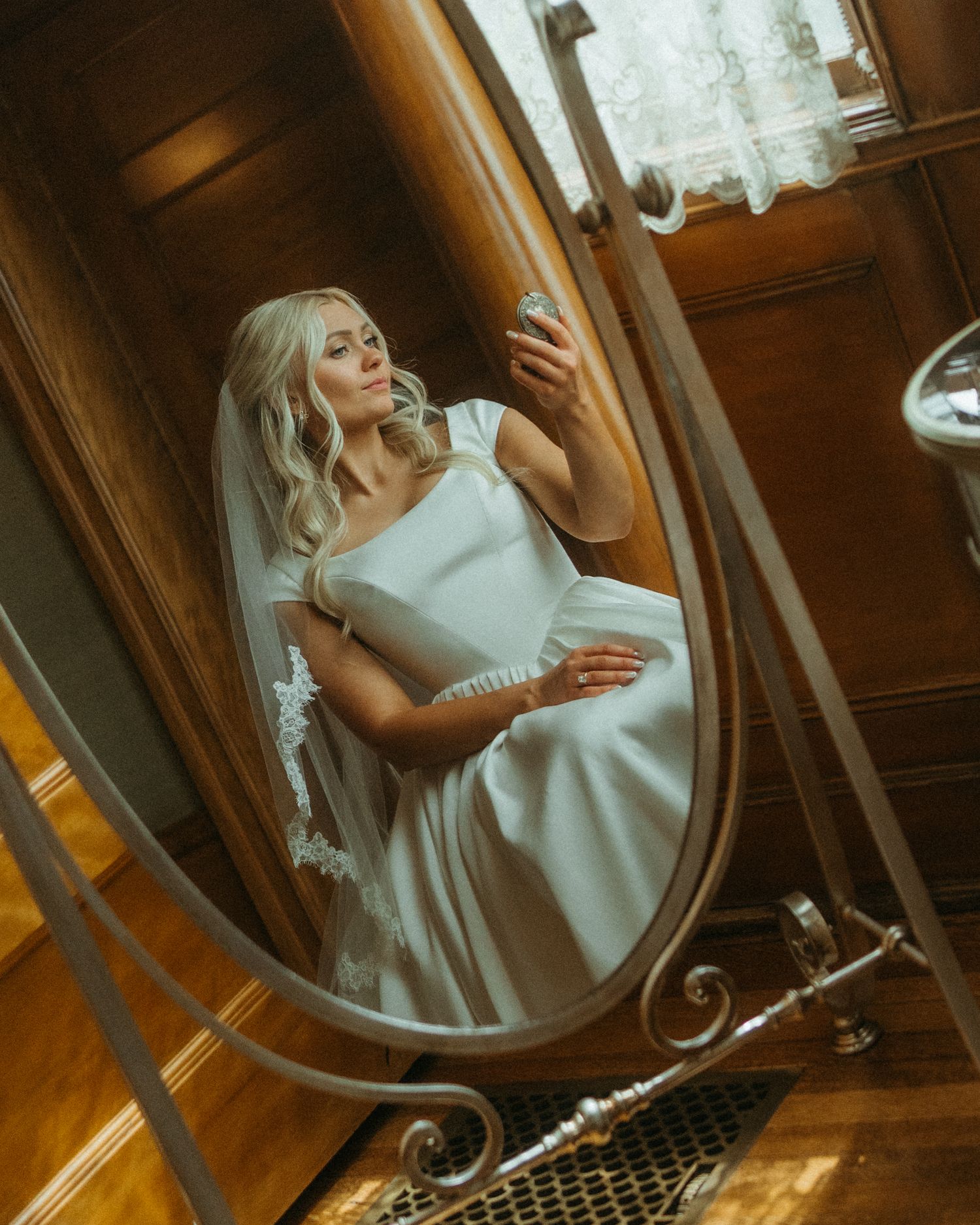 Woman in white dress gazes at her reflection in an ornate vintage mirror in warm lighting.