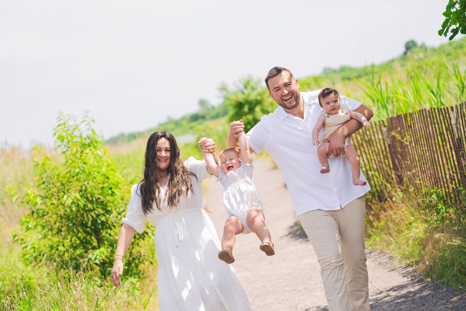A happy family in white outfits walks and plays together on a sunny path surrounded by greenery.