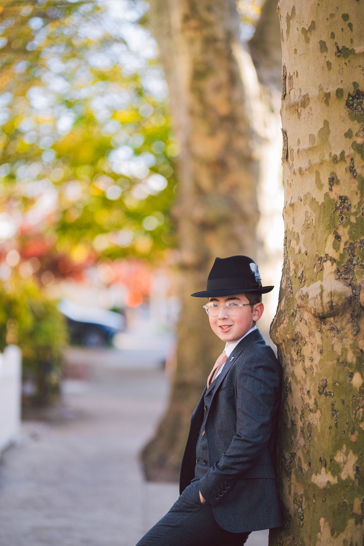 Person in black fedora and suit jacket poses playfully against tree trunk in autumn setting.