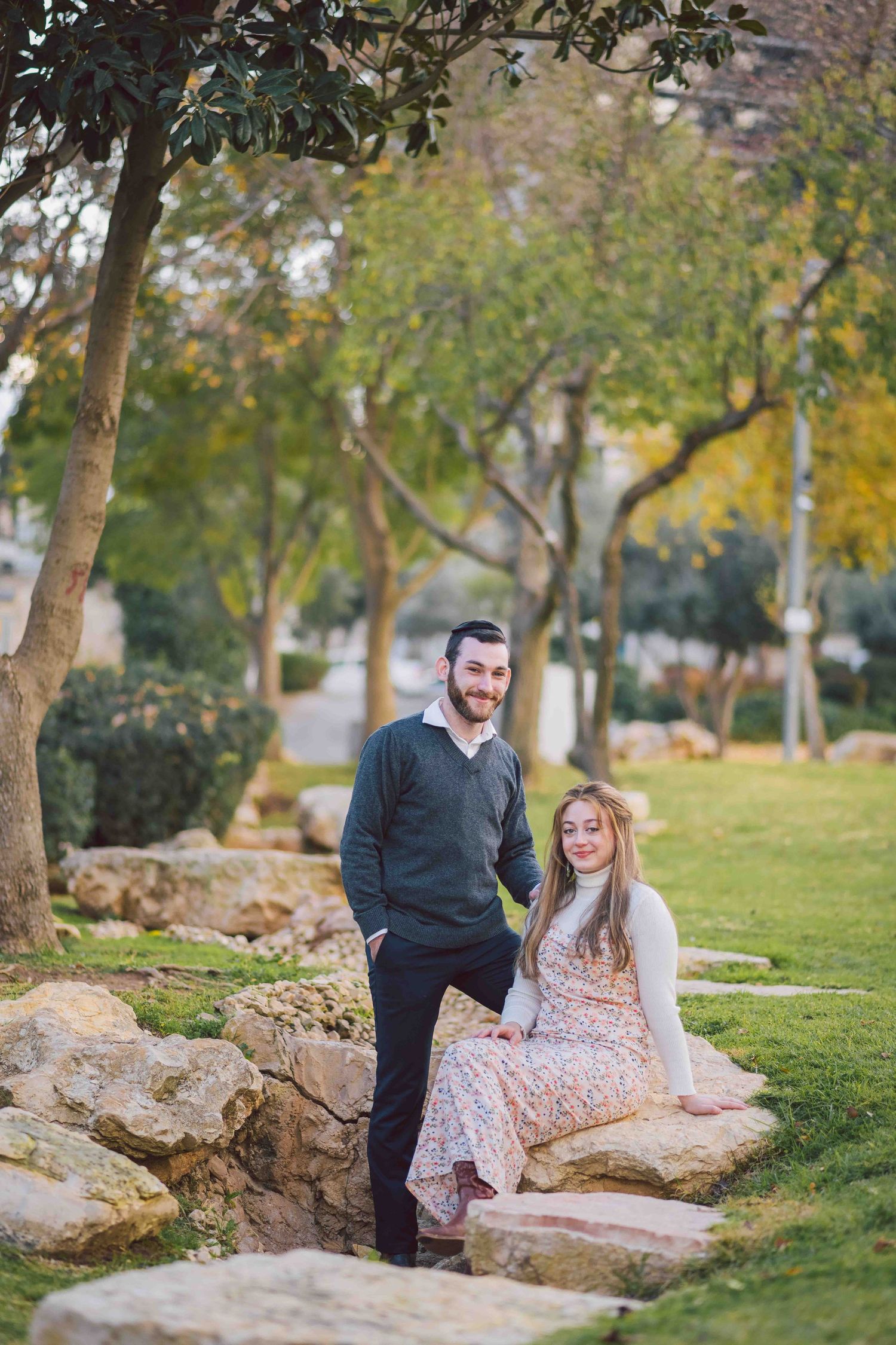 A couple sits together on large rocks in a serene park setting surrounded by trees and grass.