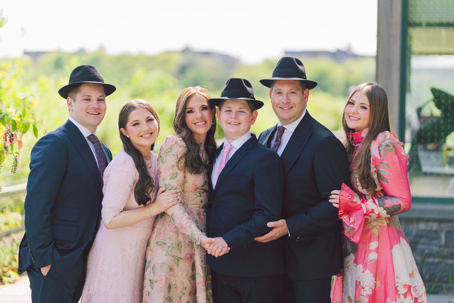 A cheerful family of six poses outdoors, dressed elegantly in pink and navy outfits with stylish black hats.