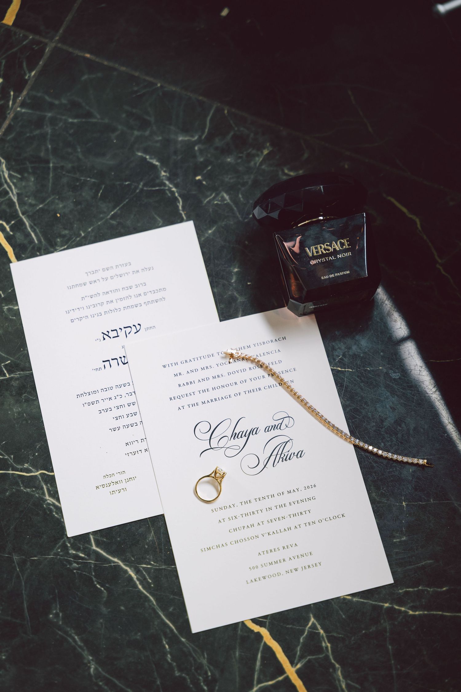 Jewish wedding invitation suite with Hebrew text, gold ring, pearl bracelet, and Versace perfume on marble surface.