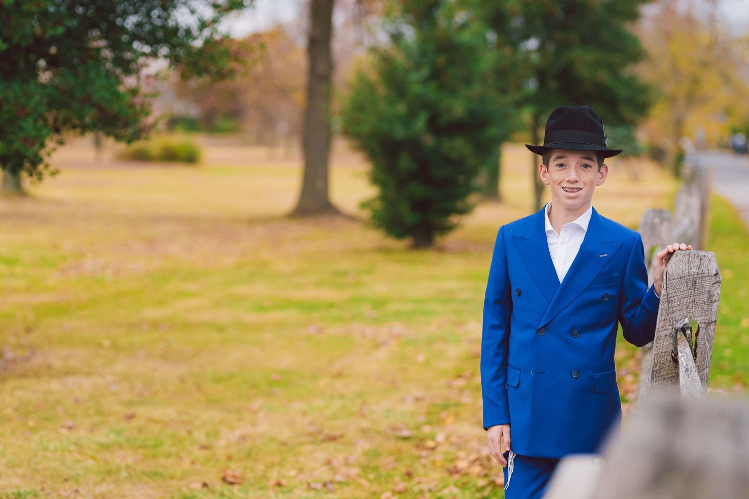 Person in bright blue suit and black hat poses near wooden post in outdoor setting.