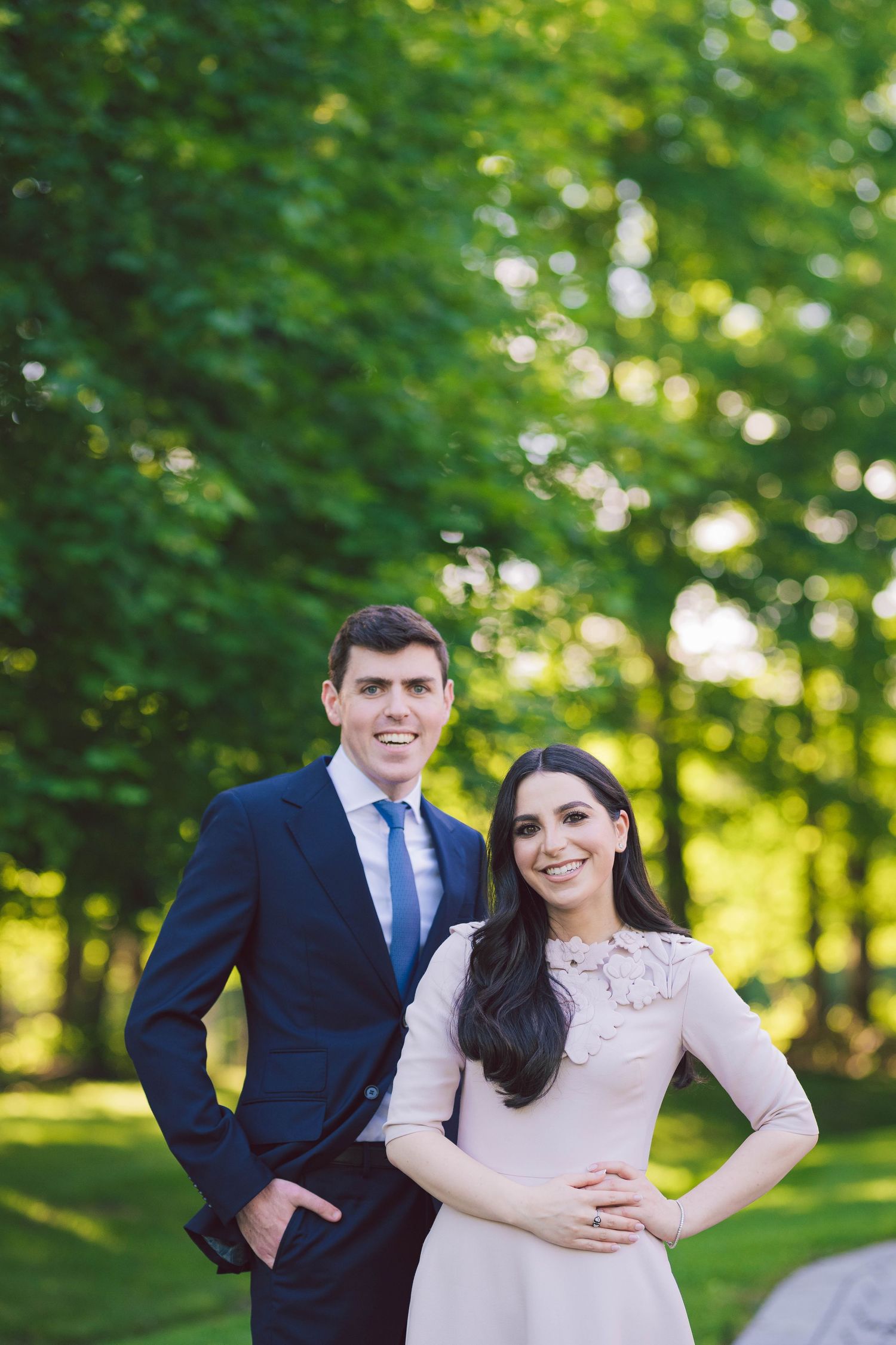 A couple poses together in formal attire against a lush green outdoor backdrop.