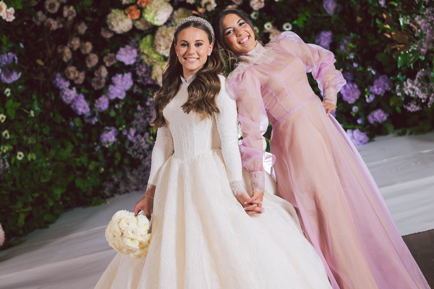 Wedding party with pink and white dresses poses by floral garden wall.