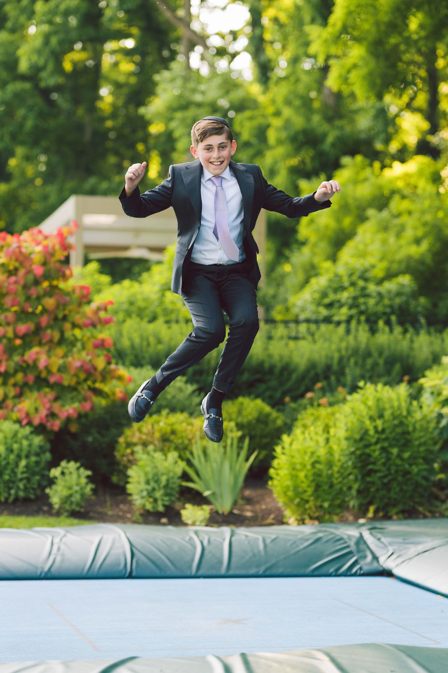 A smiling boy in a suit and tie jumps joyfully on a trampoline surrounded by lush green garden bushes.
