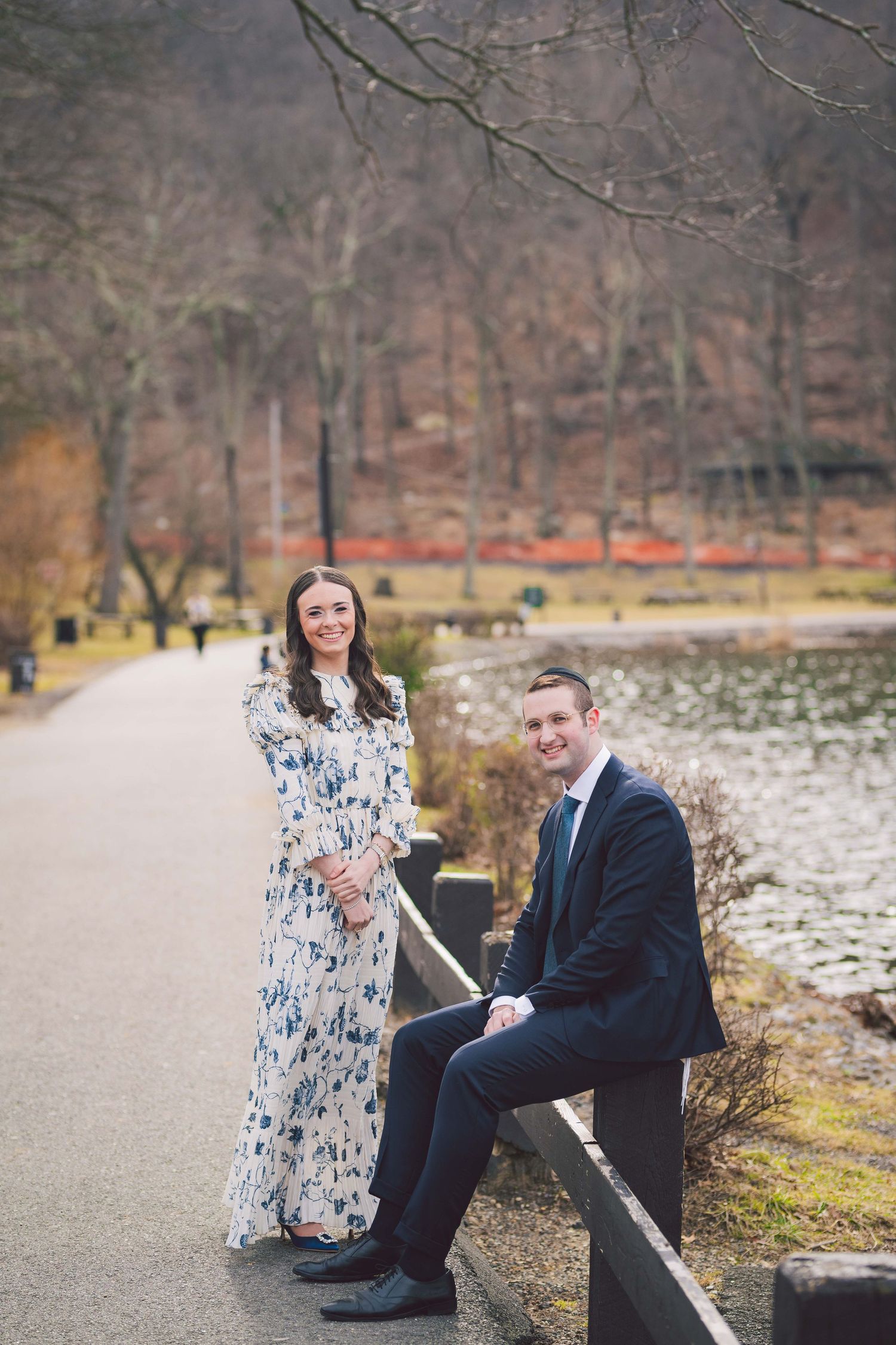 A couple sits on a wooden railing near a lake with bare winter trees in the background.