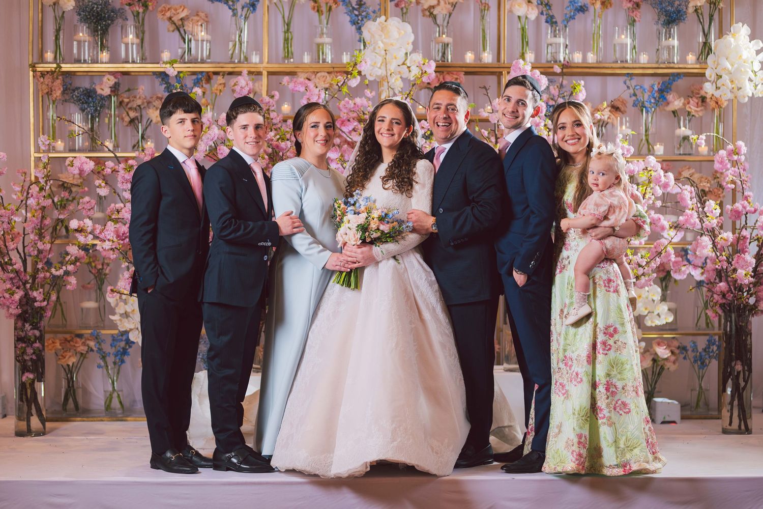 Family poses together at elegant wedding ceremony with pink floral backdrop, bride in white gown surrounded by loved ones.