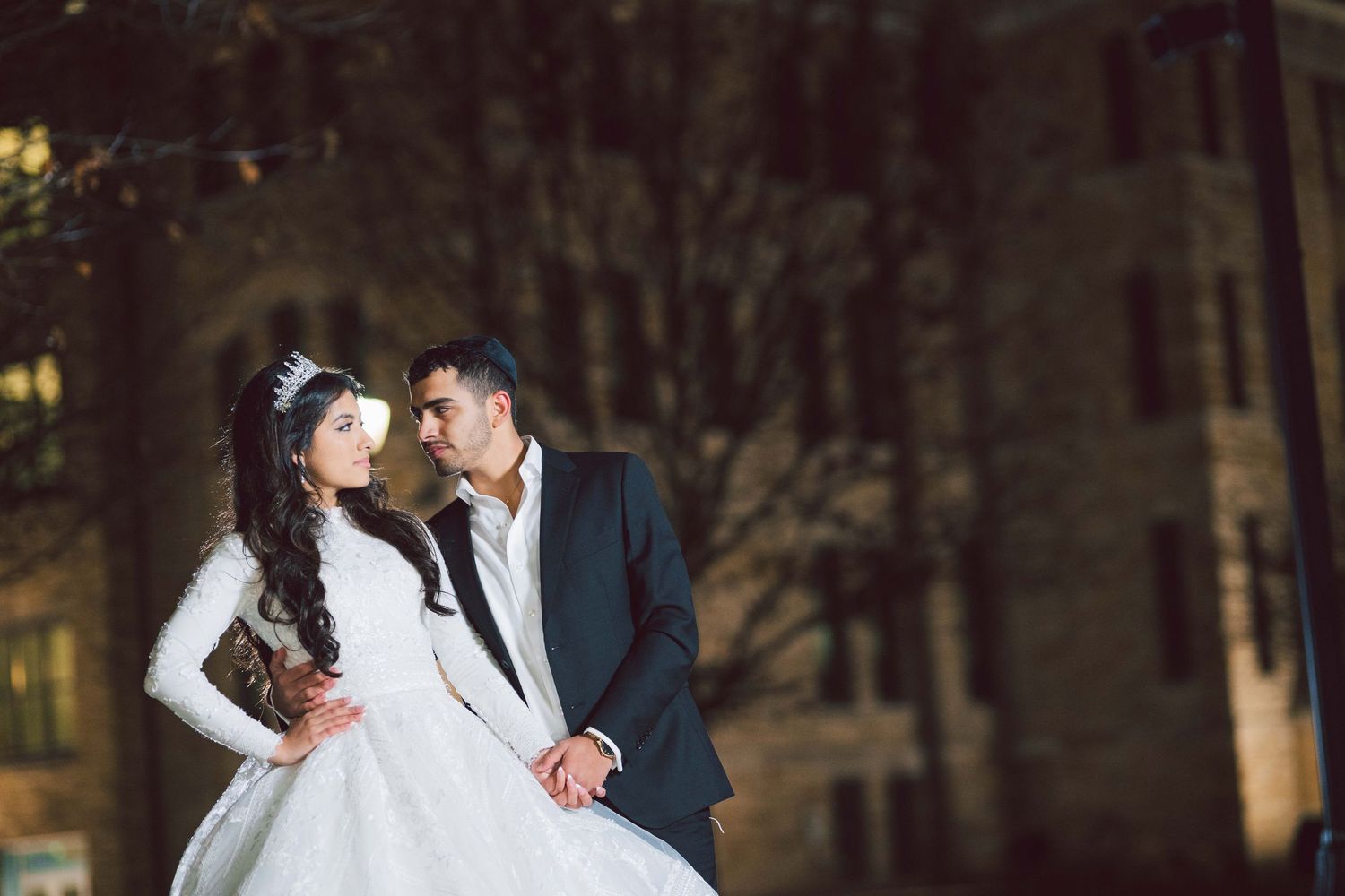Bride in white gown and groom in navy suit share a romantic moment outdoors at night during their wedding.