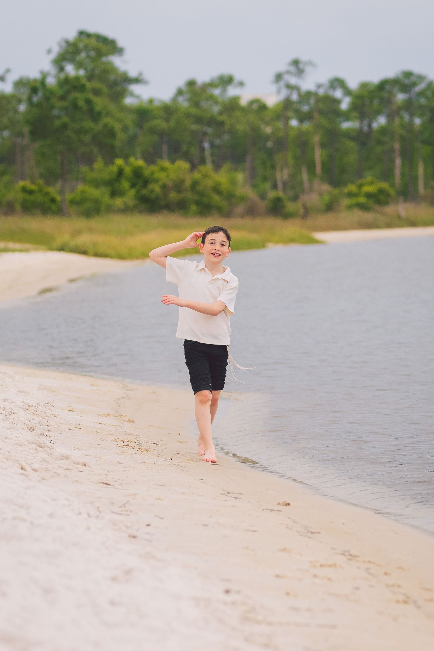 A young boy in a white shirt and black shorts runs barefoot along a sandy beach shoreline with trees in the background.