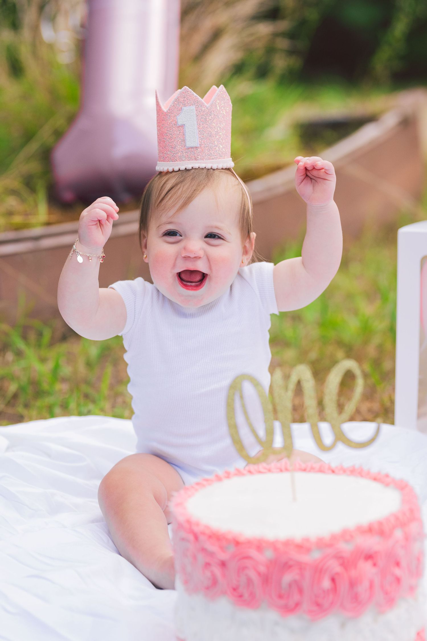 A baby in a white dress and pink crown celebrates their first birthday with a pink-frosted cake outdoors.