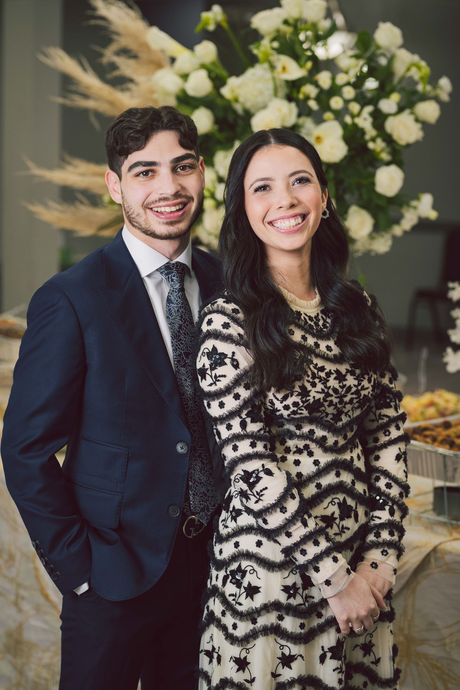 Couple poses together indoors, wearing formal attire with black and white patterned dress.