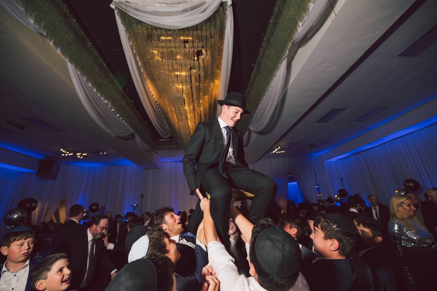 People dancing and celebrating at a wedding reception under decorative lighting and white drapery.