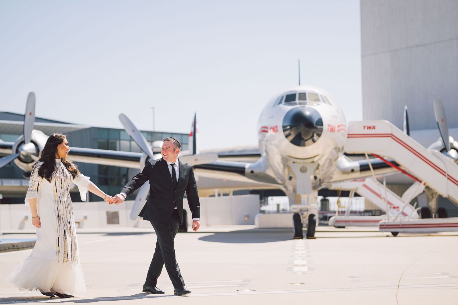 Couple walking together on airport tarmac with vintage aircraft in background.