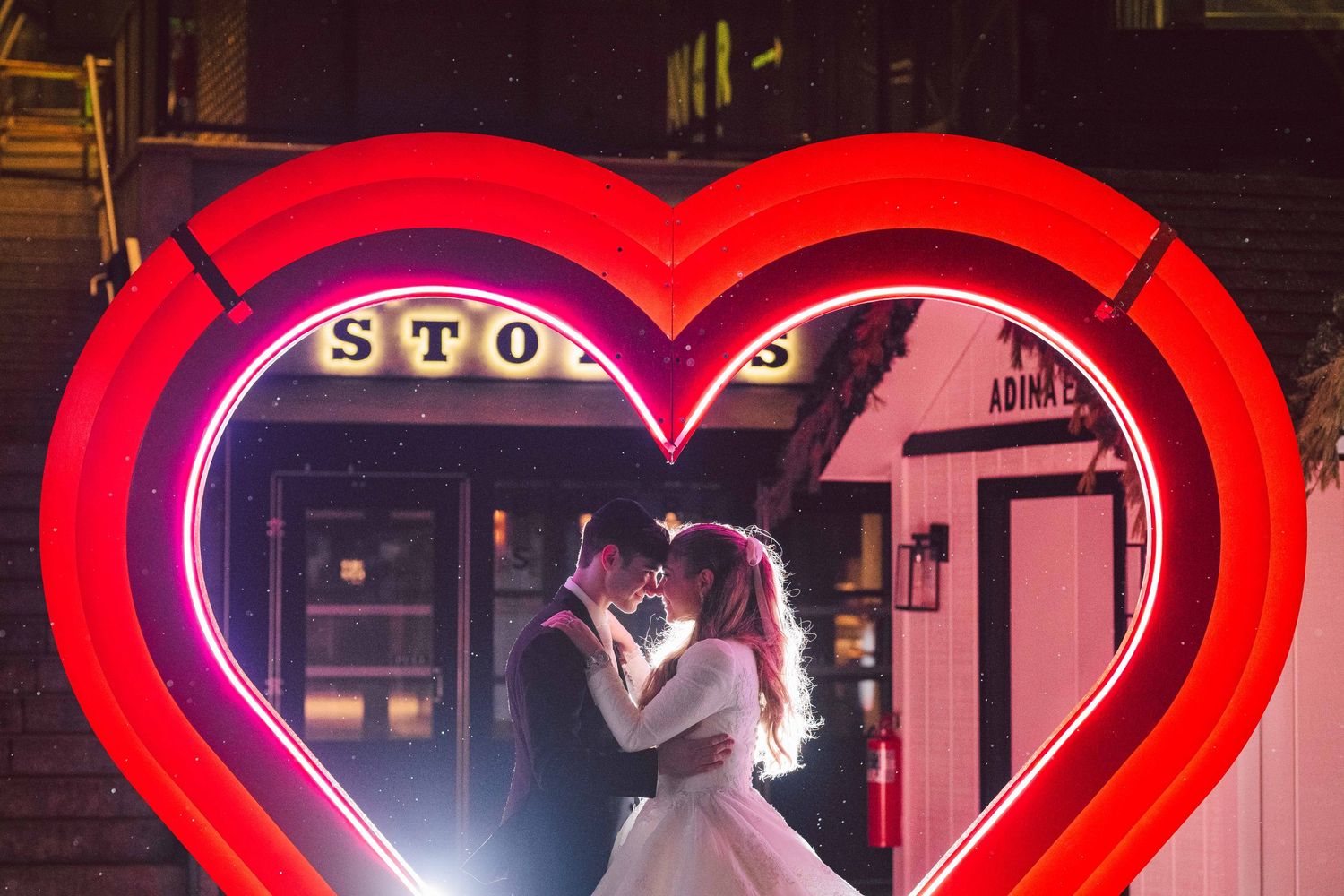 A couple in wedding attire embraces inside a glowing red neon heart.