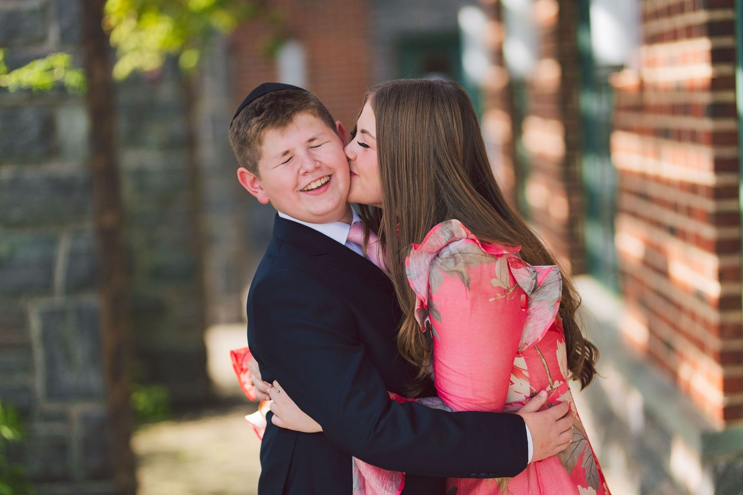 A young woman in a pink floral dress kisses a smiling boy in a dark suit outdoors near a brick building.