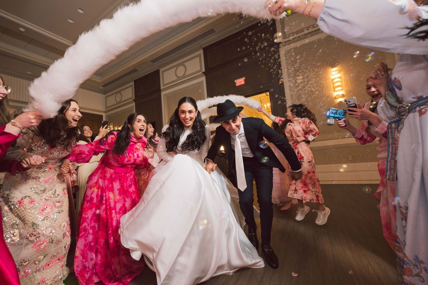 Bride and groom celebrate joyfully with guests throwing confetti and feather boa at elegant wedding reception.
