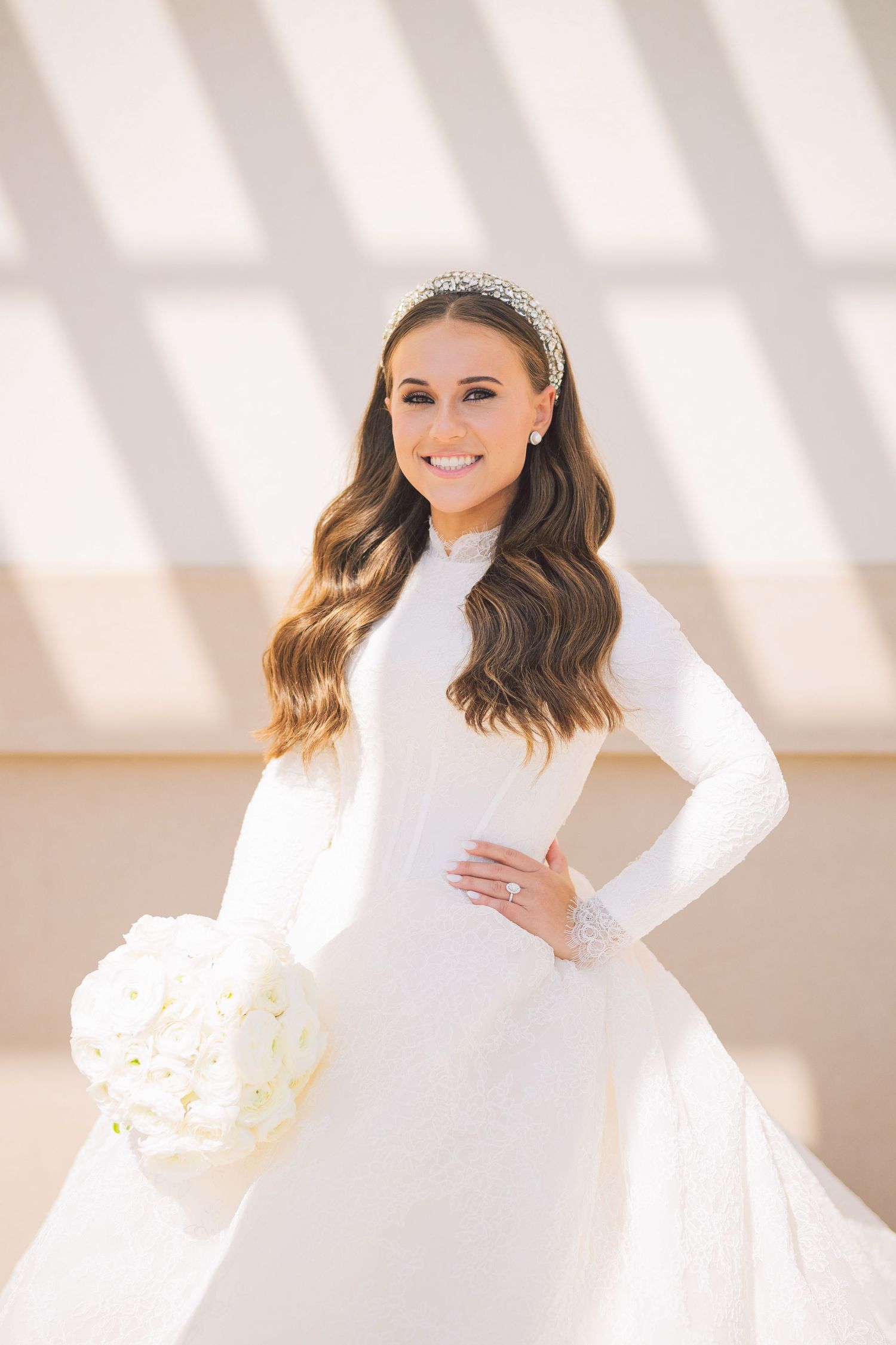 Bride in long-sleeved white wedding gown poses against sunlit geometric backdrop.