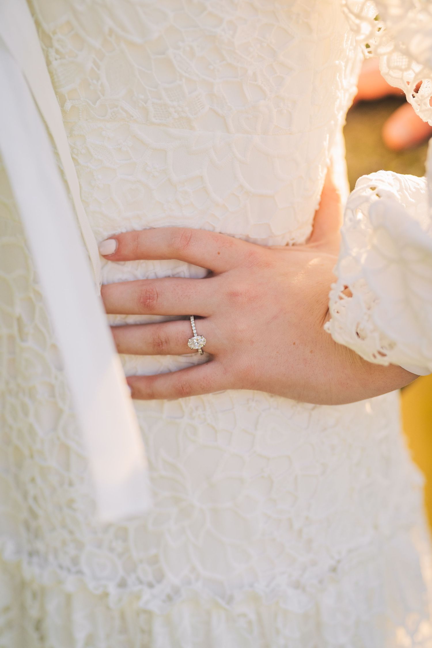 Close-up of engagement ring on hand against white lace dress.