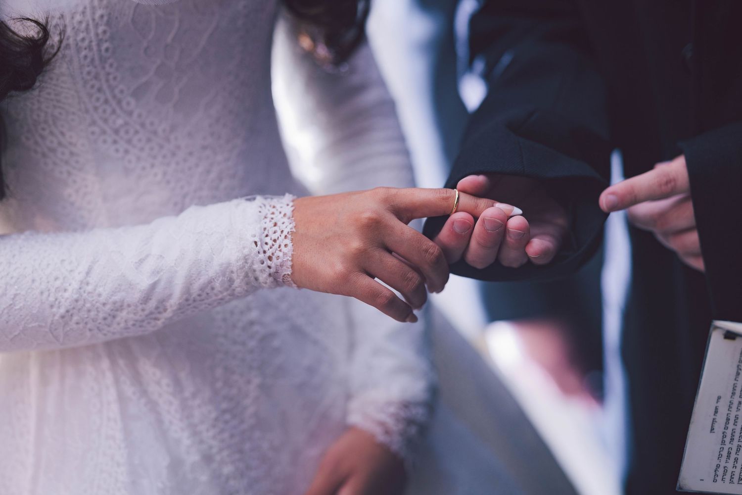 Groom placing a wedding ring on bride's finger during a ceremony, bride wearing elegant white lace dress.
