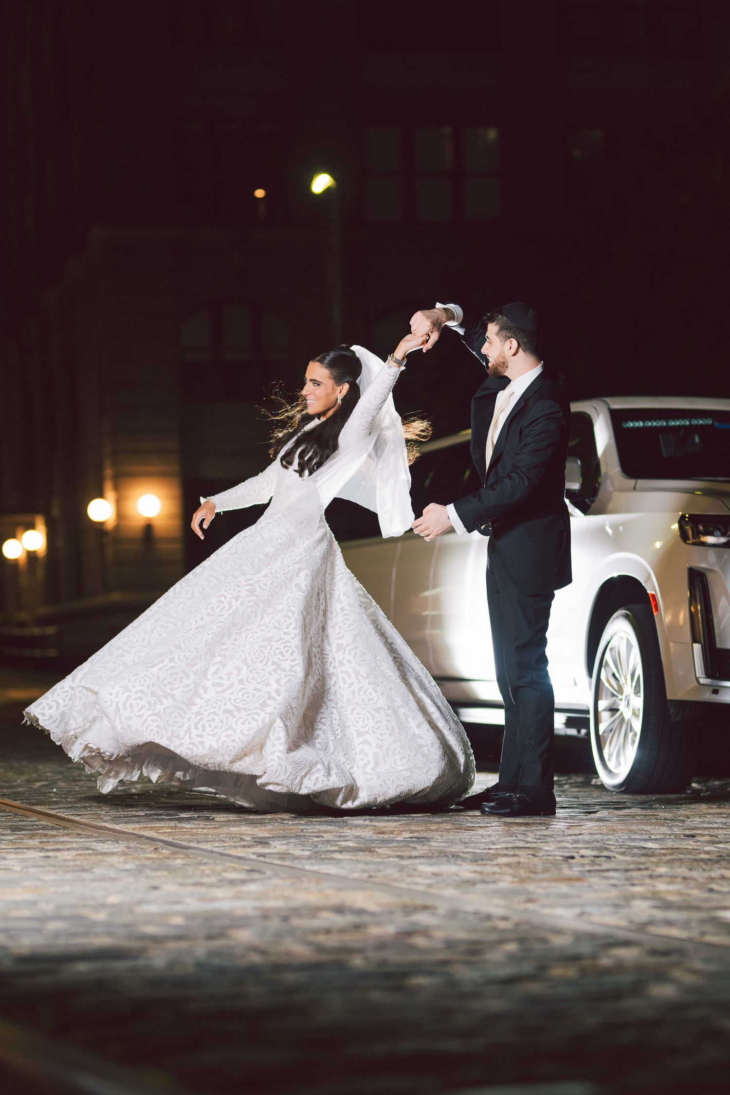 A bride in a flowing white ballgown dances with her partner on a dark cobblestone street.