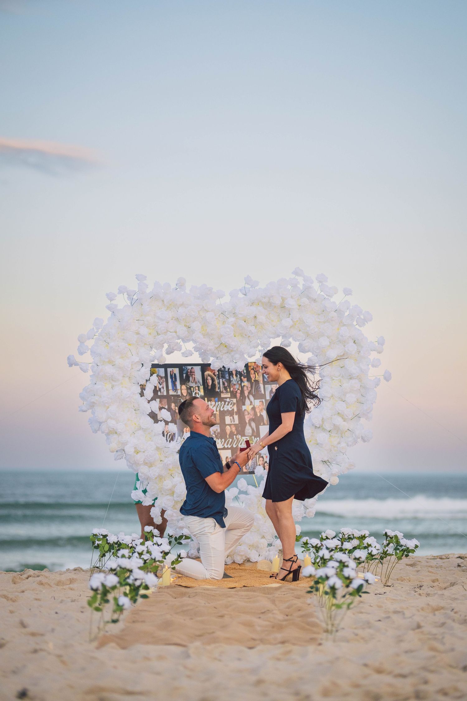 Beach proposal scene decorated with white orchid arch and ocean backdrop at sunset.