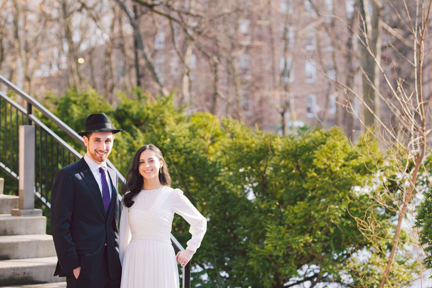 Elegant couple posing in garden setting with city buildings in background.