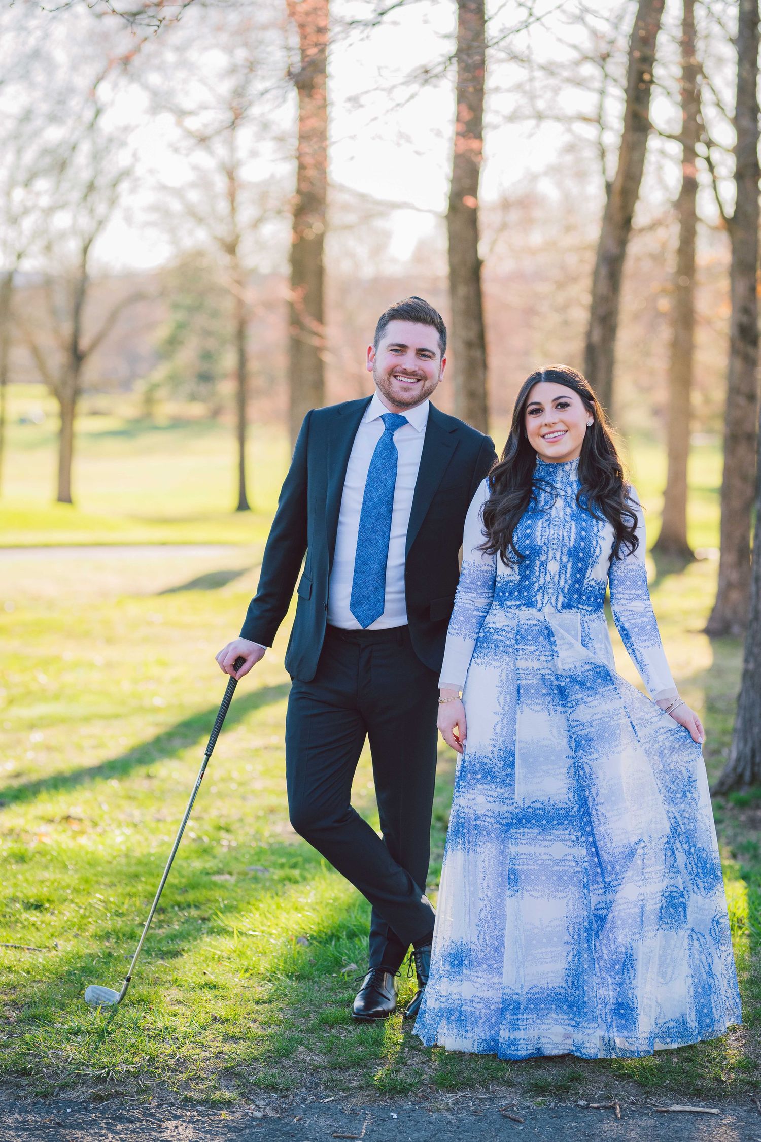 Couple in formal wear poses playfully in sunny park setting.